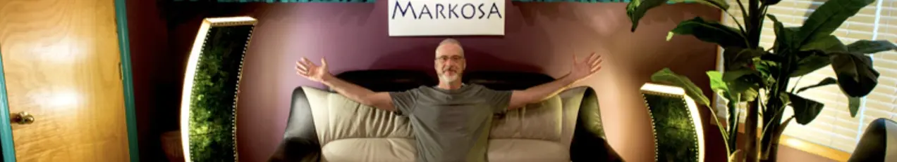 Mark Thies smiling with his arms spread wide on the couch beneath the Markosa banner